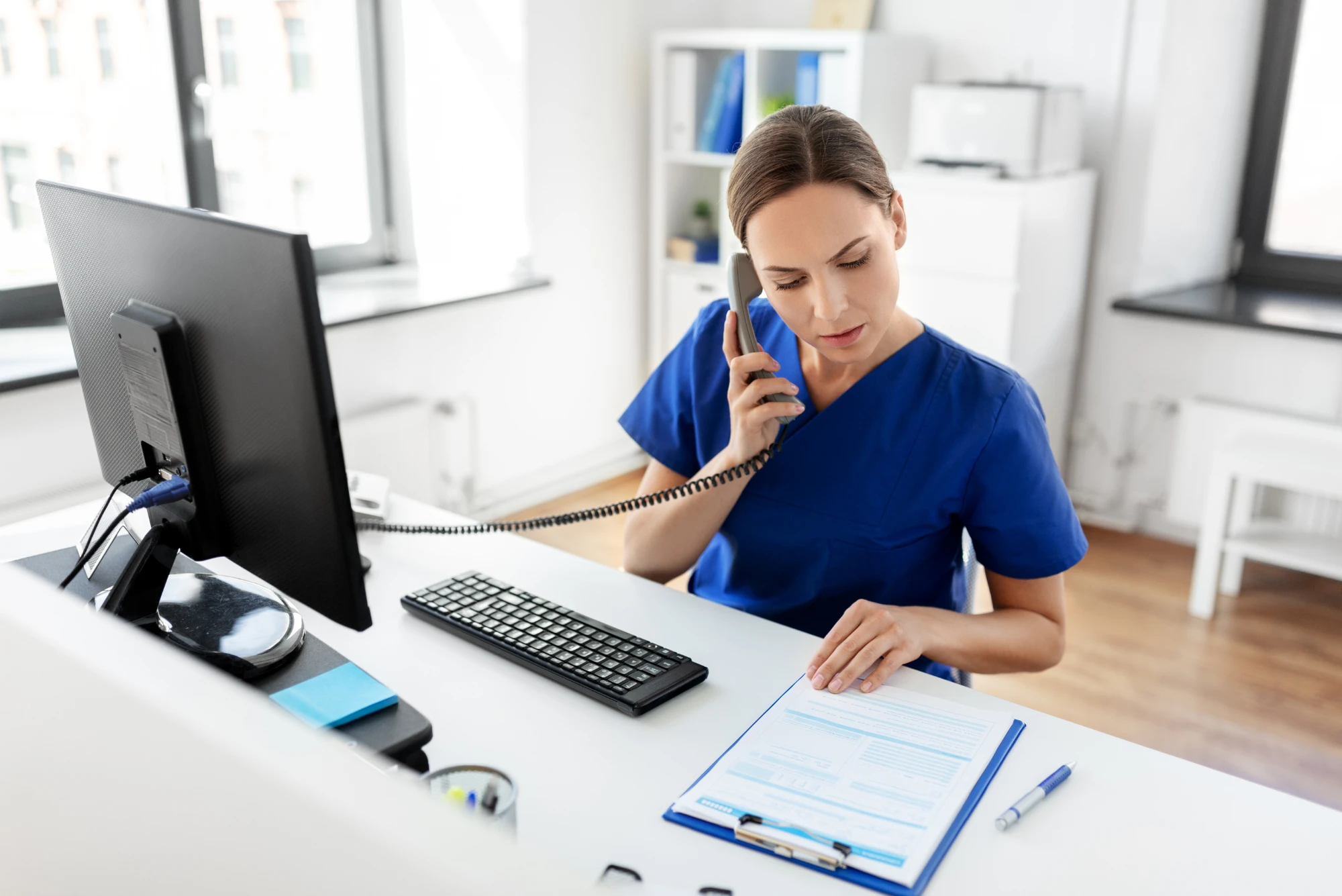 A medical front office team member is on the phone with a patient while trying to fill out paper forms because her office needs front office automation.