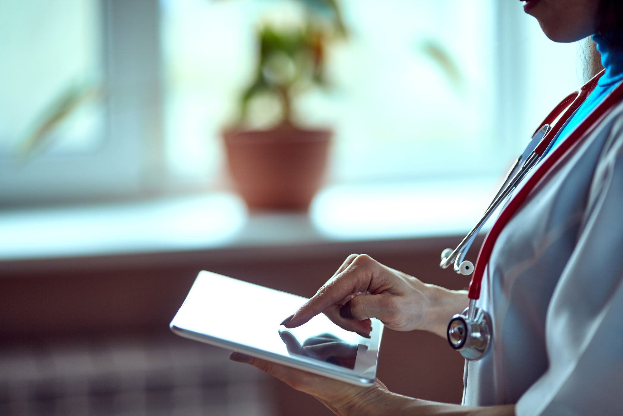 A doctor works on an iPad in a dimly-lit room with a red stethoscope around her neck.