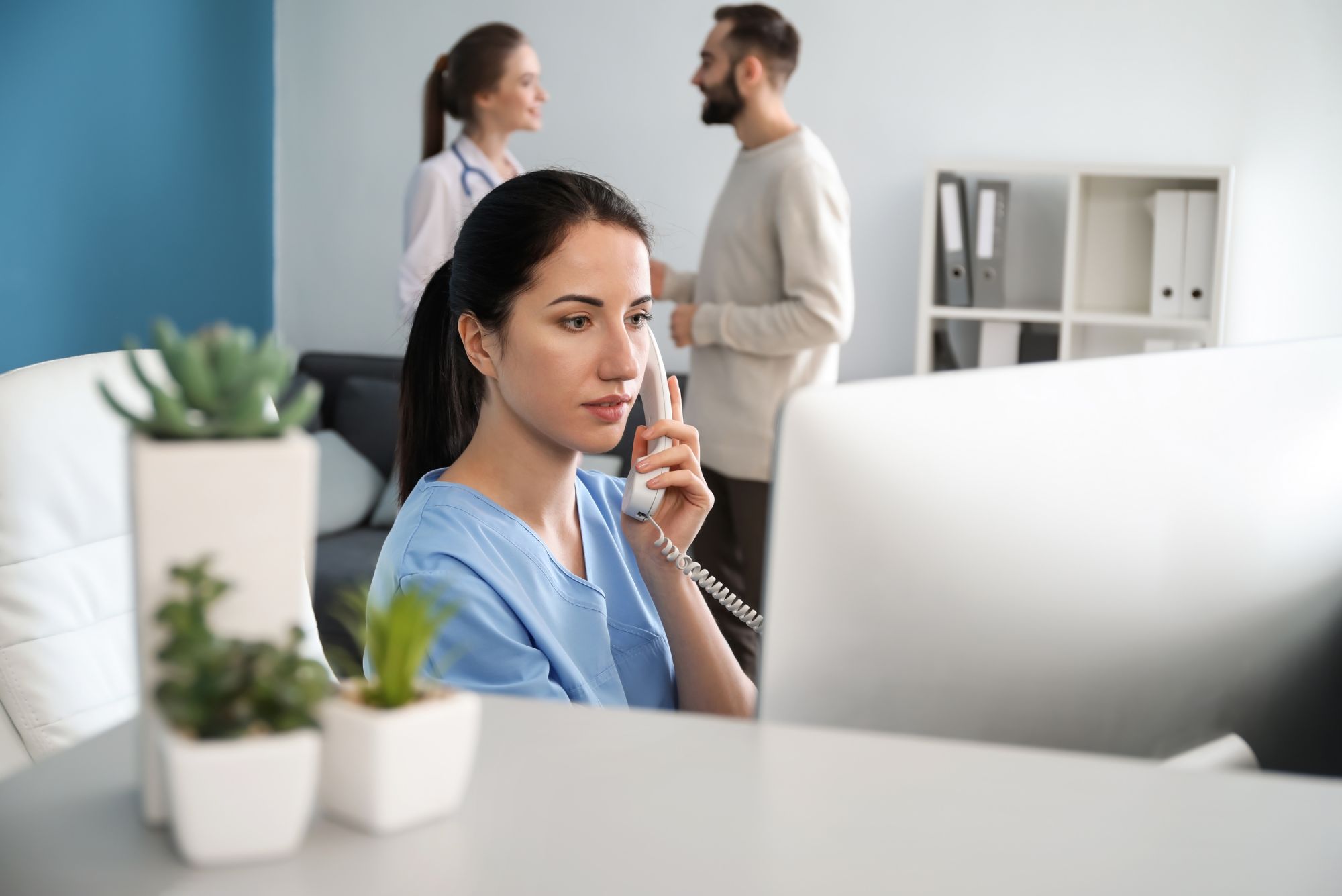 A woman sits at her desk in a medical office, using front office automation while taking patient calls.