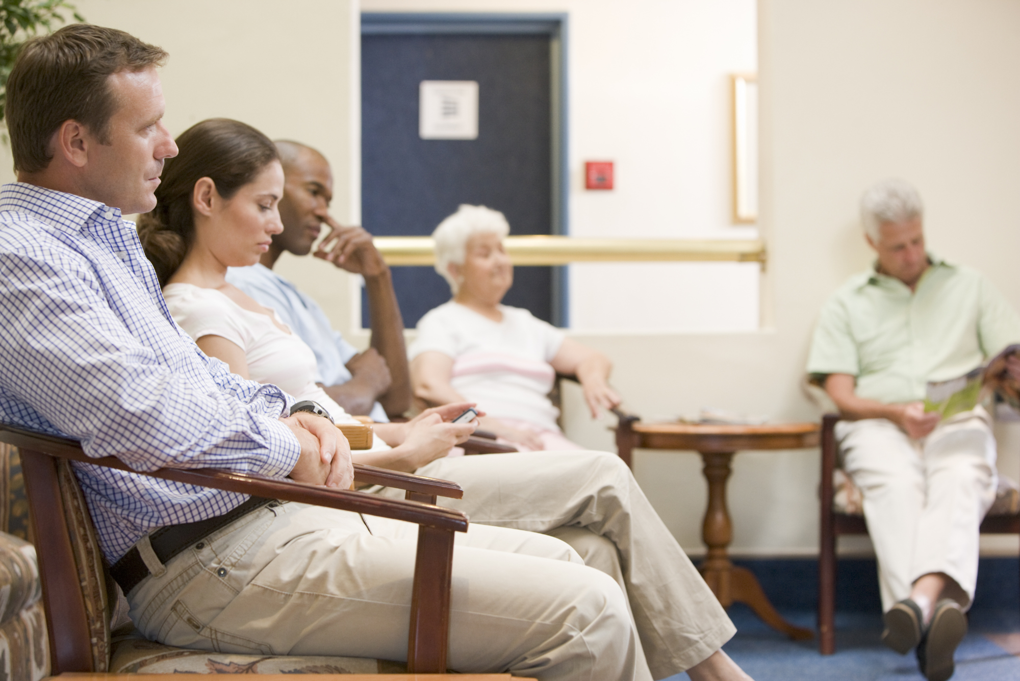 Five people sit in chairs in a waiting room because their doctor’s office needs a patient engagement solution for the waiting room.